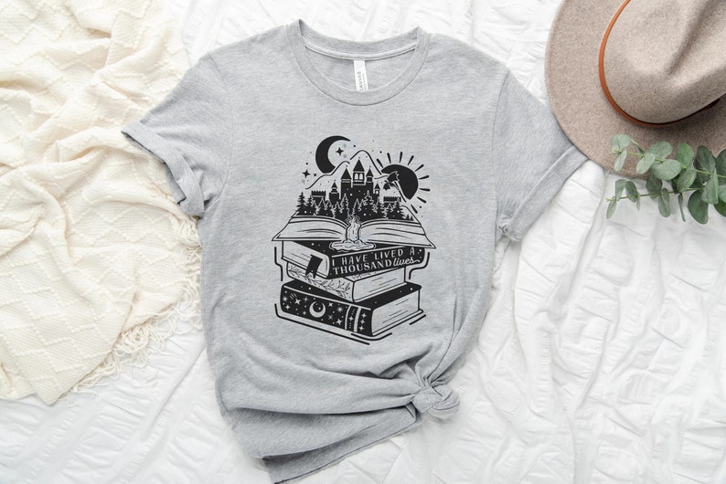 May include: A gray t-shirt with a black and white graphic of a stack of books with a castle and forest scene on the top book. The text on the shirt reads "I have lived a thousand lives."