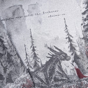 May include: A light grey sweatshirt featuring a forest and dragon design. The design includes a dragon, trees, and the text "from now until the darkness claims us." The cuffs and hem are a solid light grey.