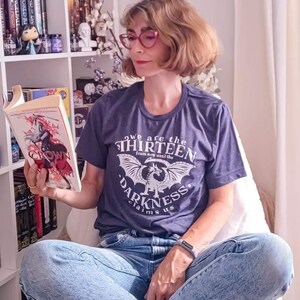 May include: A woman wearing a navy blue t-shirt with the text "we are the thirteen from now until the darkness claims us" and light blue jeans, reading a book titled "Crown of Midnight". The background includes a bookshelf with books and decorative items.