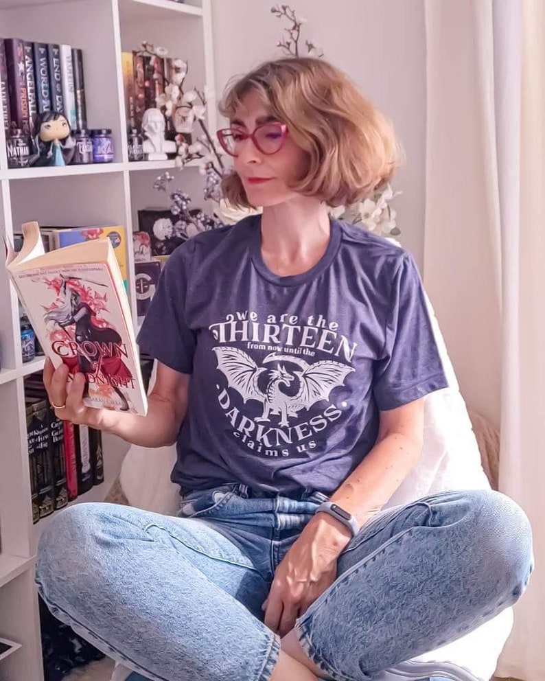 May include: A woman wearing a navy blue t-shirt with the text "we are the thirteen from now until the darkness claims us" and light blue jeans, reading a book titled "Crown of Midnight". The background includes a bookshelf with books and decorative items.