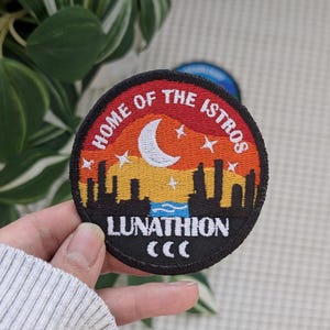 May include: Embroidered patch with a circular design featuring a cityscape silhouette under a sunset sky. The text reads "HOME OF THE ISTROS" and "LUNATHION" with a crescent moon and stars.