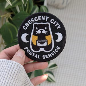 May include: Black circular patch with the text "CRESCENT CITY POSTAL SERVICE". The patch features a cartoon animal holding a letter, with yellow and white accents. The patch is held in a hand.