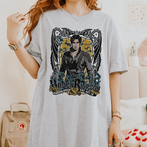 May include: Grey t-shirt featuring a graphic print of a man with a dragon design. The text on the shirt reads "Hayden Riderson, Riderson House, Sat Oct 10".