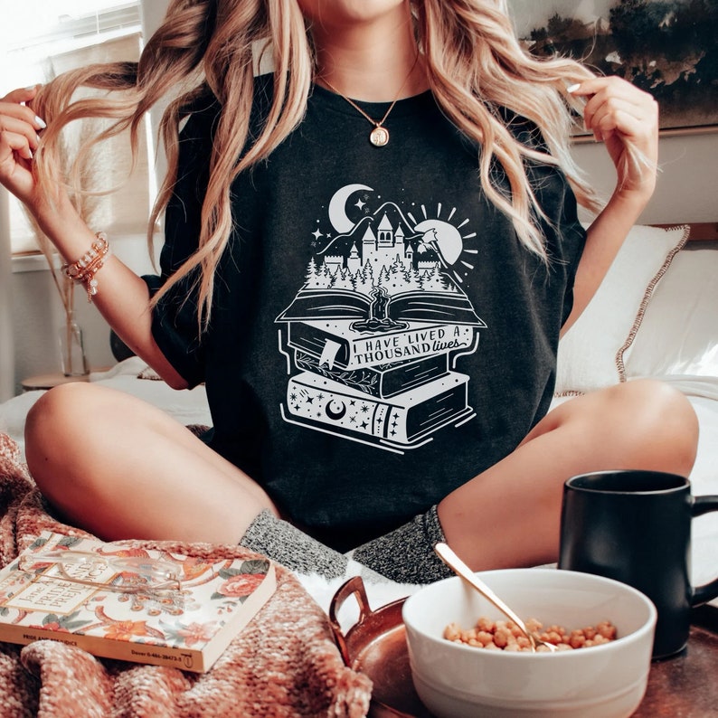 May include: Black t-shirt with a white graphic of a stack of books with a castle and forest scene on top. The text "I have lived a thousand lives" is printed on the books.
