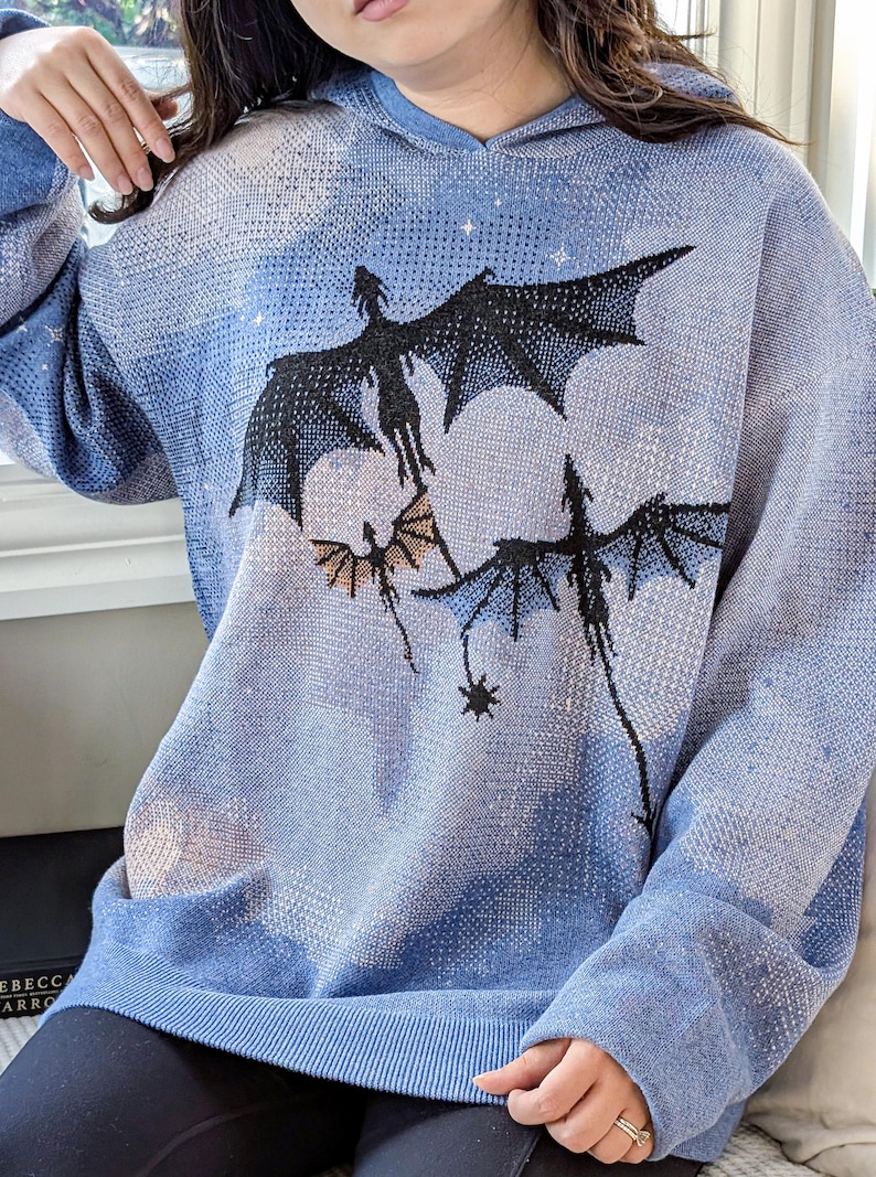 K&ouml;nnte beinhalten: Ein hellblauer Strick-Kapuzenpullover mit einem eingewebten Grafikmuster aus drei schwarzen Drachen, die vor einem bew&ouml;lkten Himmel mit kleinen wei&szlig;en Sternen fliegen. Der lang&auml;rmelige Pullover hat einen gerippten Saum, B&uuml;ndchen und eine Kapuze.