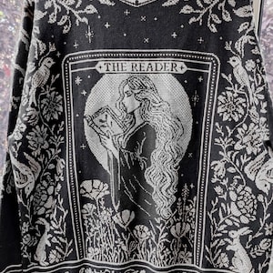 May include: Black knitted sweater featuring a white illustration of a woman reading, with floral and rabbit patterns. The text "THE READER" is displayed above the image. The sweater has a crew neck and ribbed trims.