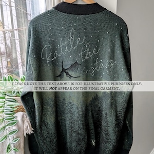 May include: A dark green long-sleeved sweater with a black collar and a white design of a dragon flying through a starry sky. The text "Rattle the stars" is written in white above the dragon.