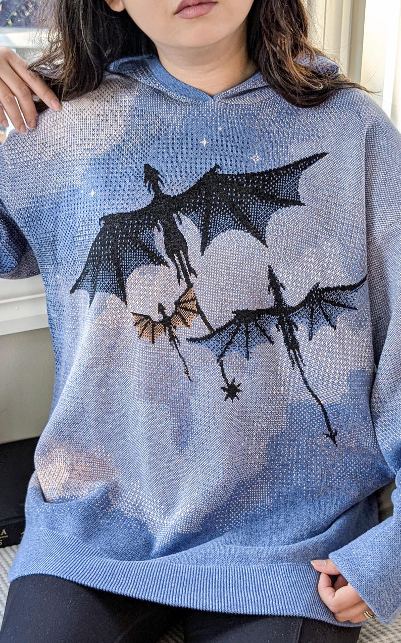 K&ouml;nnte beinhalten: Ein hellblauer Strick-Kapuzenpullover mit einem grafischen Muster aus drei schwarzen Drachen, die vor einem himmelsartigen Hintergrund mit wei&szlig;en Wolken und kleinen Sternen fliegen. Das Kleidungsst&uuml;ck hat lange &Auml;rmel und einen gerippten Saum.