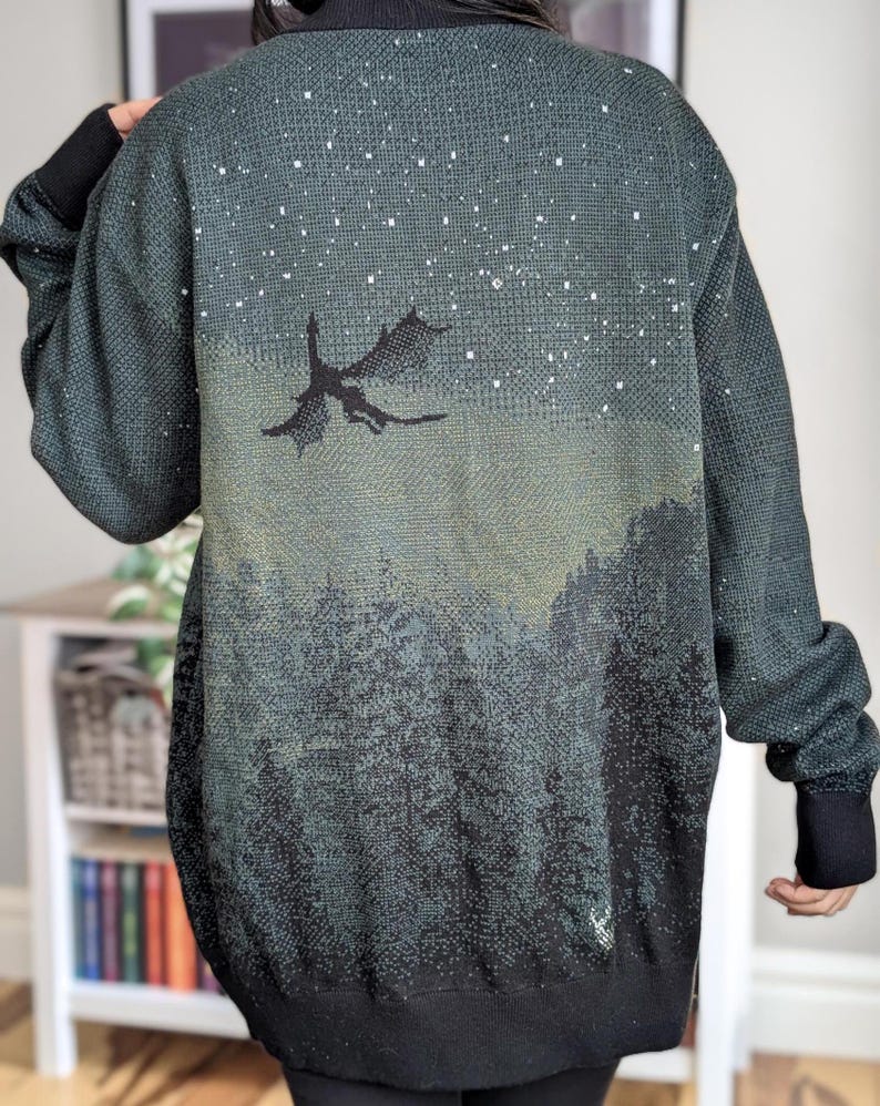 May include: A dark green cardigan with a black silhouette of a dragon flying against a starry night sky. The cardigan has a black knit collar and cuffs.