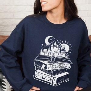 May include: Navy blue sweatshirt with a white graphic of a castle, trees, and a crescent moon on top of a stack of books. The text "I have lived a thousand lives" is printed on the books.