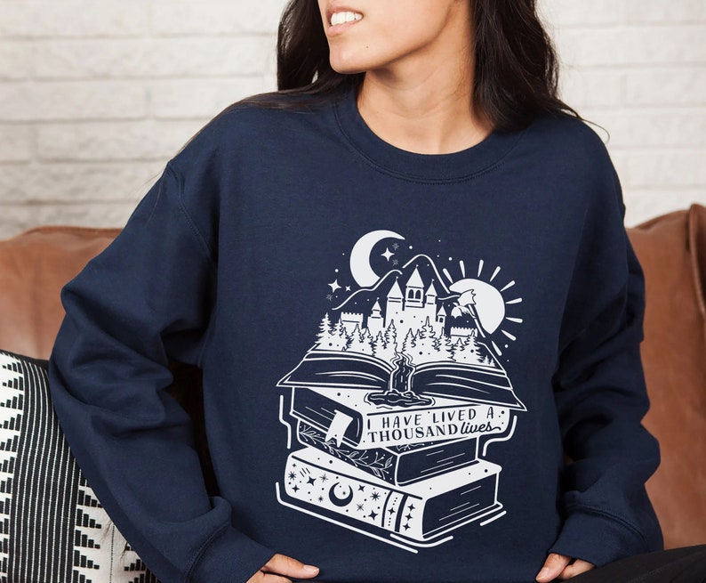 May include: Navy blue sweatshirt with a white graphic of a castle, trees, and a crescent moon on top of a stack of books. The text "I have lived a thousand lives" is printed on the books.