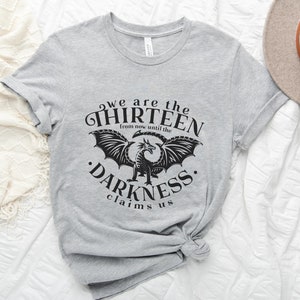 May include: A light gray t-shirt with the text "we are the THIRTEEN from now until the DARKNESS claims us" in black. A black dragon graphic is centered below the text. A beige hat and greenery are in the background.