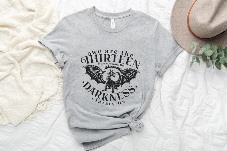 May include: A light gray t-shirt with the text "we are the THIRTEEN from now until the DARKNESS claims us" in black. A black dragon graphic is centered below the text. A beige hat and greenery are in the background.