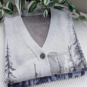 May include: A folded gray cardigan sweater with a V-neck and a single button closure. The sweater features a landscape design with trees in shades of gray and black. A patterned blue and purple fabric is visible beneath the sweater.