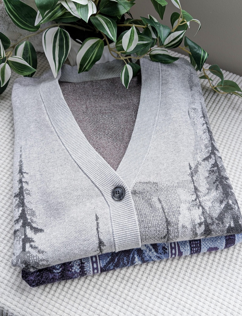 May include: A folded gray cardigan sweater with a V-neck and a single button closure. The sweater features a landscape design with trees in shades of gray and black. A patterned blue and purple fabric is visible beneath the sweater.