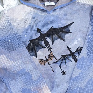 K&ouml;nnte beinhalten: Ein blauer Kapuzenpullover mit einem Strickmuster aus Wolken und drei fliegenden schwarzen Drachen. Das Design enth&auml;lt kleine wei&szlig;e Sternakzente, die &uuml;ber den himmelsartigen Hintergrund verteilt sind. Das Kleidungsst&uuml;ck hat lange &Auml;rmel und eine Kapuze.