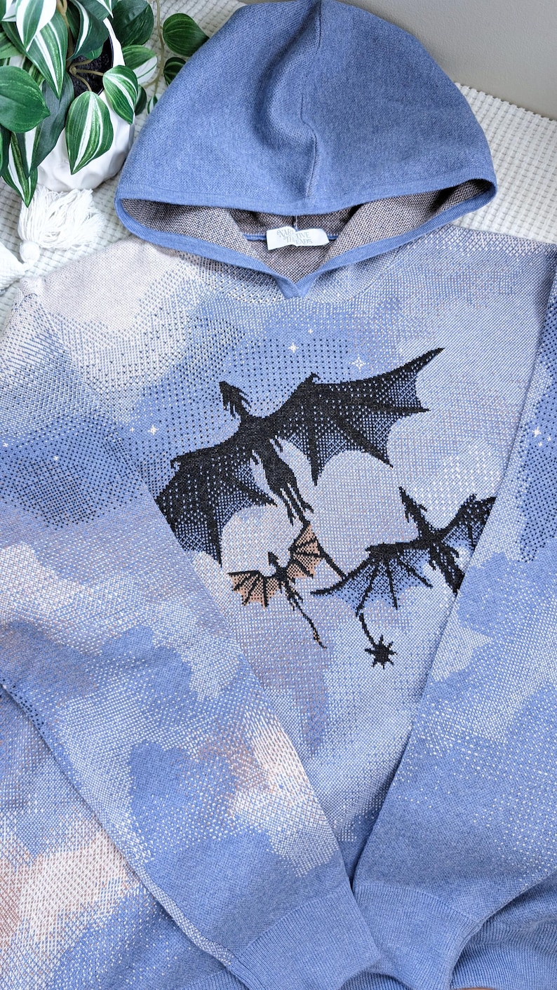 K&ouml;nnte beinhalten: Ein blauer Kapuzenpullover mit einem Strickmuster aus Wolken und drei fliegenden schwarzen Drachen. Das Design enth&auml;lt kleine wei&szlig;e Sternakzente, die &uuml;ber den himmelsartigen Hintergrund verteilt sind. Das Kleidungsst&uuml;ck hat lange &Auml;rmel und eine Kapuze.