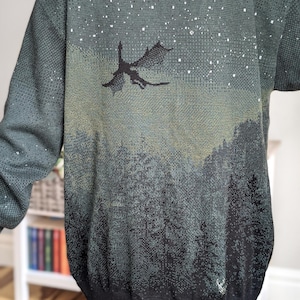 May include: A dark green knitted cardigan with a gradient design featuring a silhouette of a dragon flying over a forest scene. The cardigan has a speckled pattern of white dots throughout.