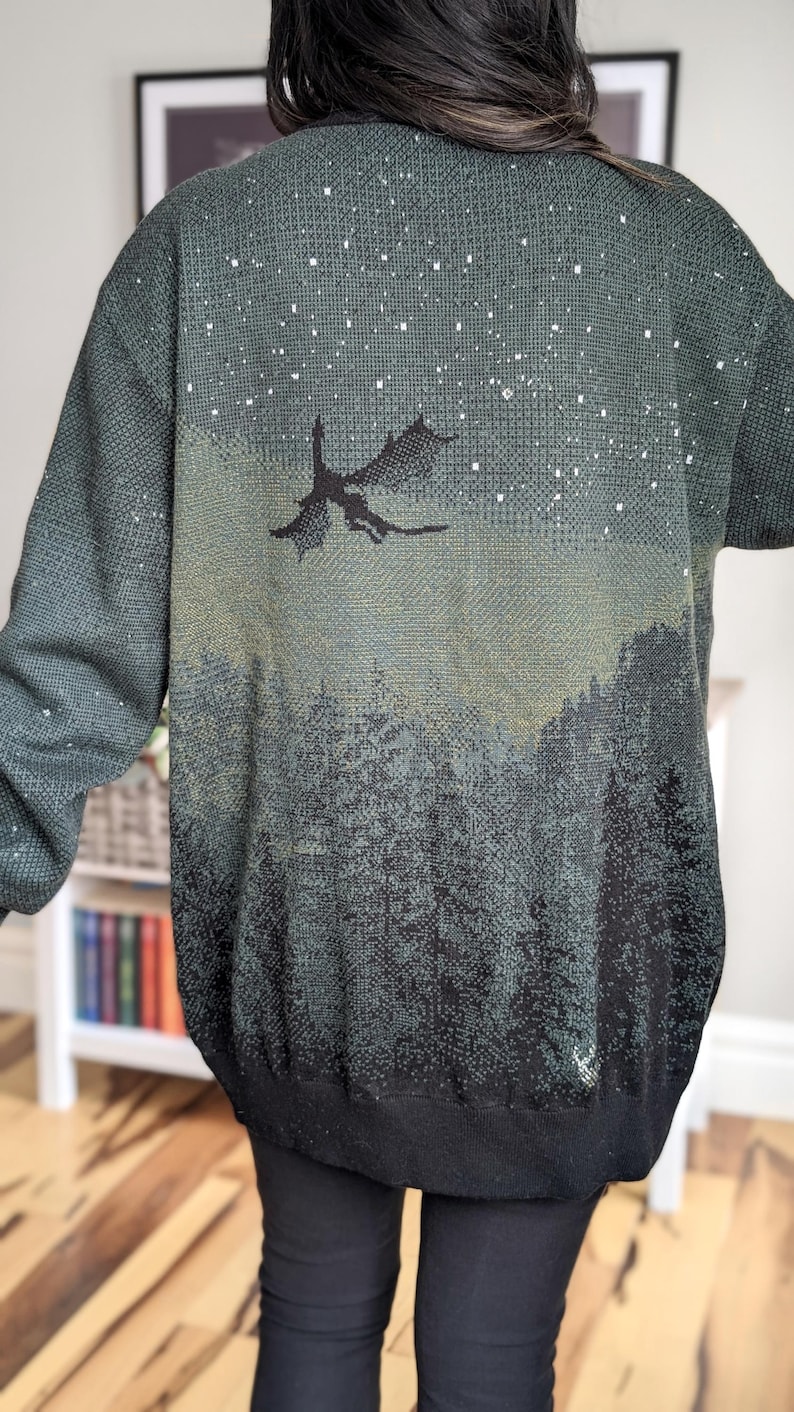 May include: A dark green knitted cardigan with a gradient design featuring a silhouette of a dragon flying over a forest scene. The cardigan has a speckled pattern of white dots throughout.