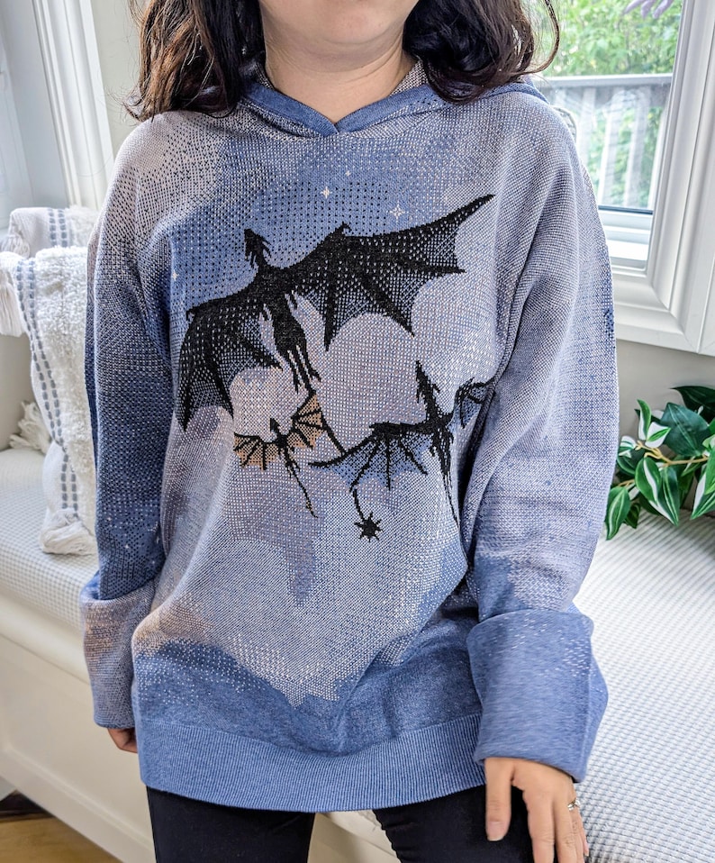 K&ouml;nnte beinhalten: Ein blauer Kapuzenpullover mit einem gestrickten Grafikmuster aus drei fliegenden Drachen in Schwarz und Gold vor einem melierten blau-wei&szlig;en Himmelshintergrund. Das Kleidungsst&uuml;ck hat lange &Auml;rmel, eine Kapuze und einen gerippten Saum.