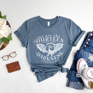May include: Blue t-shirt with the text "we are the thirteen from now until the darkness claims us" and a dragon graphic. The shirt is paired with blue jeans and white sneakers. A wooden tray with flowers and accessories is also in the image.