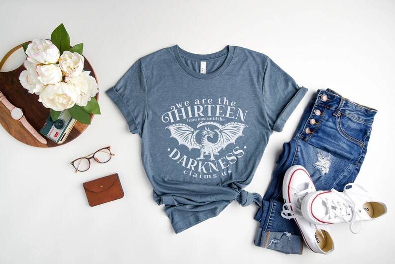 May include: Blue t-shirt with the text "we are the thirteen from now until the darkness claims us" and a dragon graphic. The shirt is paired with blue jeans and white sneakers. A wooden tray with flowers and accessories is also in the image.