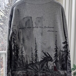 May include: Gray sweater with a forest and dragon design. The back of the sweater features a dark forest scene with a dragon and a figure in a red cloak. The text "from now until the darkness claims us" is printed on the back.