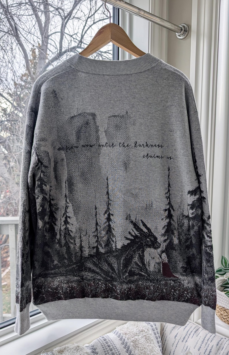 May include: Gray sweater with a forest and dragon design. The back of the sweater features a dark forest scene with a dragon and a figure in a red cloak. The text "from now until the darkness claims us" is printed on the back.