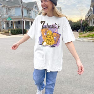 May include: A white t-shirt with a graphic of a cat sitting on a card with the text "Zihnal's Adoption Agency" printed on the front. The cat is orange and white with green eyes. The card is purple with yellow stars and a crescent moon. The text is in brown.