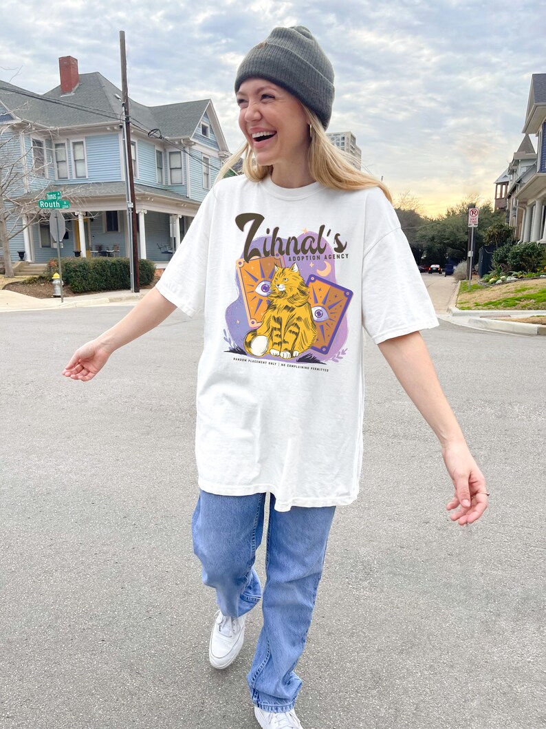 May include: A white t-shirt with a graphic of a cat sitting on a card with the text "Zihnal's Adoption Agency" printed on the front. The cat is orange and white with green eyes. The card is purple with yellow stars and a crescent moon. The text is in brown.
