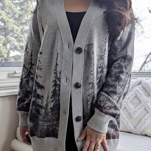 May include: A gray cardigan with a forest scene design. The button-down sweater features a V-neck and long sleeves. The forest scene is printed in black and gray, with a hint of burgundy at the bottom.