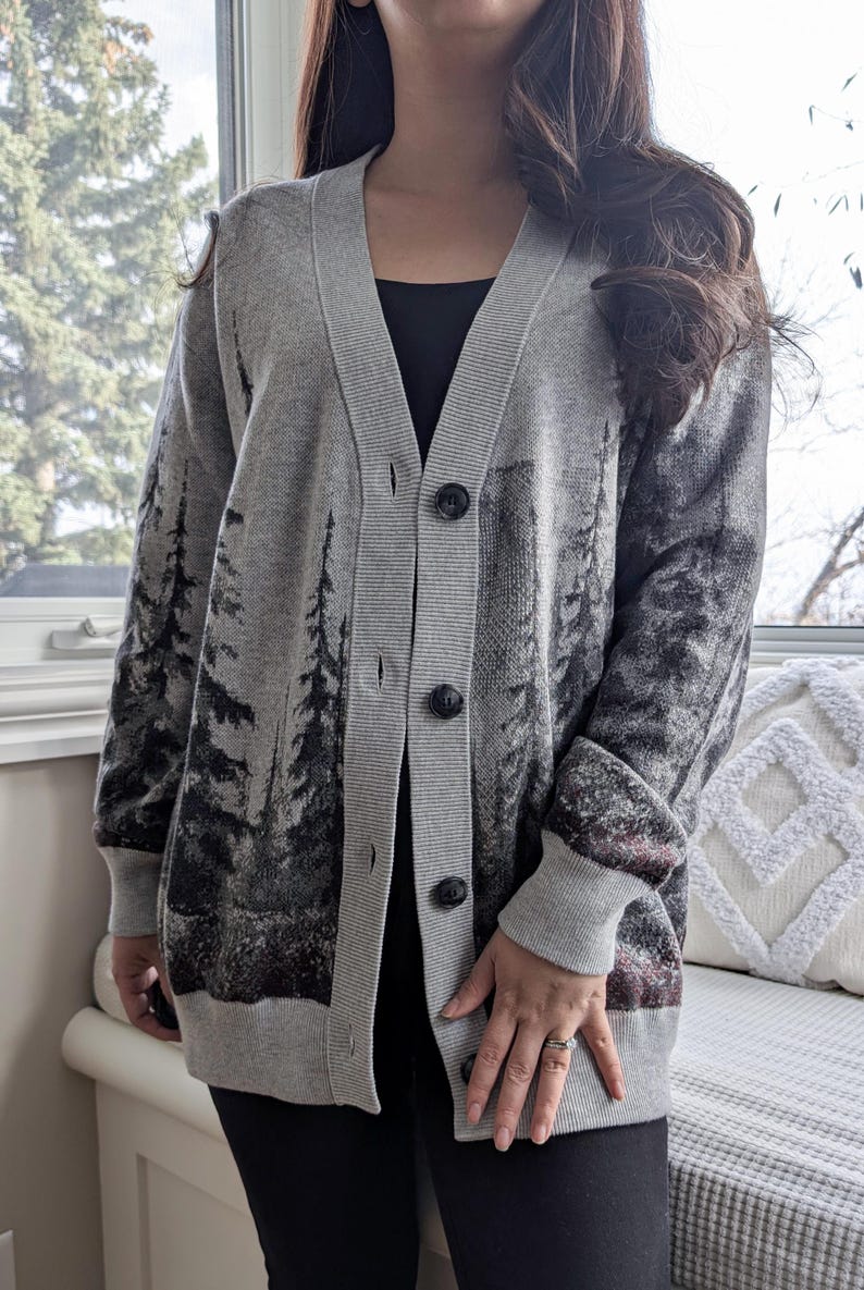 May include: A gray cardigan with a forest scene design. The button-down sweater features a V-neck and long sleeves. The forest scene is printed in black and gray, with a hint of burgundy at the bottom.