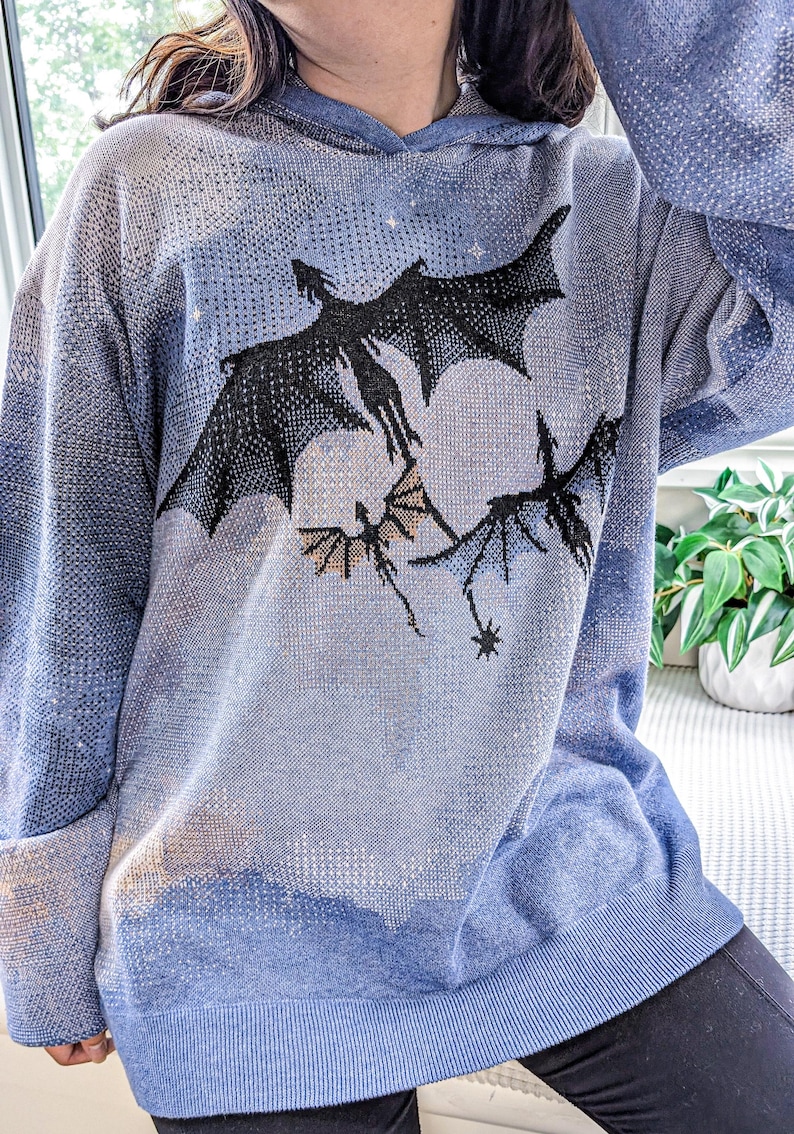 K&ouml;nnte beinhalten: Ein blauer Strick-Kapuzenpullover mit einer Grafik von drei schwarzen Drachen, die vor einem gro&szlig;en, blassen, kreisf&ouml;rmigen Mond fliegen. Der Pullover hat eine texturierte, pixelige Optik mit Farbverl&auml;ufen in Blau, langen &Auml;rmeln und einem gerippten Saum.