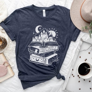 May include: A navy blue t-shirt with a white graphic of a stack of books with a castle and trees on top. The text on the book reads "I have lived a thousand lives".