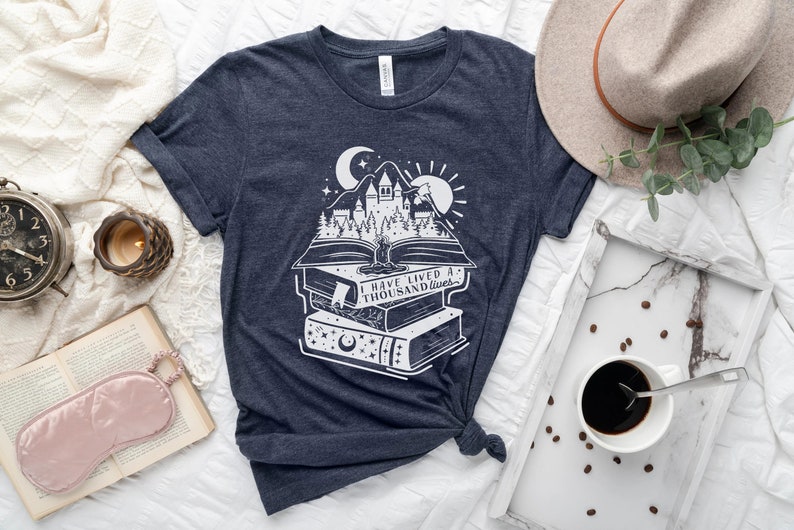 May include: A navy blue t-shirt with a white graphic of a stack of books with a castle and trees on top. The text on the book reads "I have lived a thousand lives".
