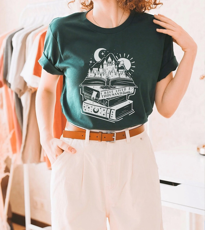May include: A dark green t-shirt with a white graphic of a castle on a stack of books. The graphic includes a crescent moon, a sun, and the text "I have lived a thousand lives."