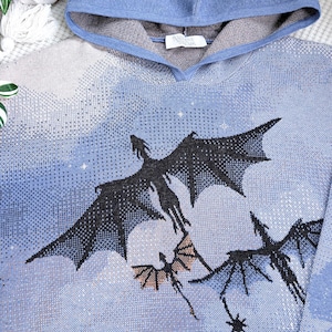 K&ouml;nnte beinhalten: Ein blauer Strick-Kapuzenpullover mit einem eingewebten Motiv von drei schwarzen Drachen, die vor einem hellblauen und wei&szlig;en bew&ouml;lkten Himmel fliegen. Die Innenseite der Kapuze ist grau abgesetzt und ein wei&szlig;es Etikett mit der Aufschrift Narrative Threads ist zu sehen.