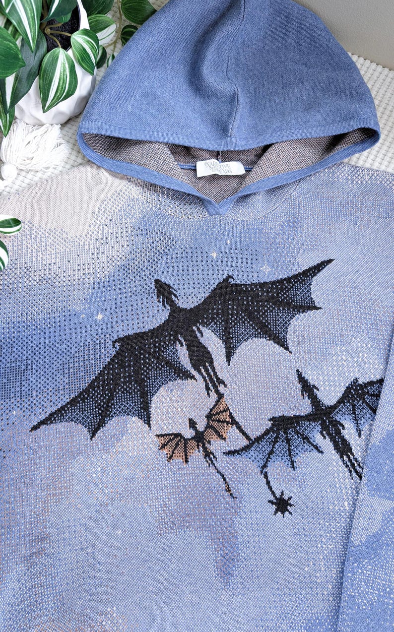K&ouml;nnte beinhalten: Ein blauer Strick-Kapuzenpullover mit einem eingewebten Motiv von drei schwarzen Drachen, die vor einem hellblauen und wei&szlig;en bew&ouml;lkten Himmel fliegen. Die Innenseite der Kapuze ist grau abgesetzt und ein wei&szlig;es Etikett mit der Aufschrift Narrative Threads ist zu sehen.