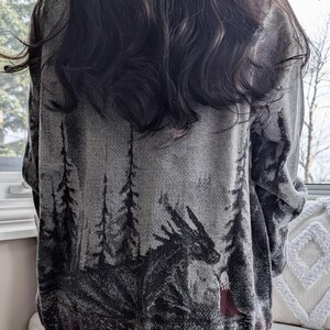 May include: A gray sweater featuring a forest scene with a dragon and a figure in red. The design is in shades of gray and black, with a gray hem and cuffs. The sweater is worn by a person with long, dark hair.