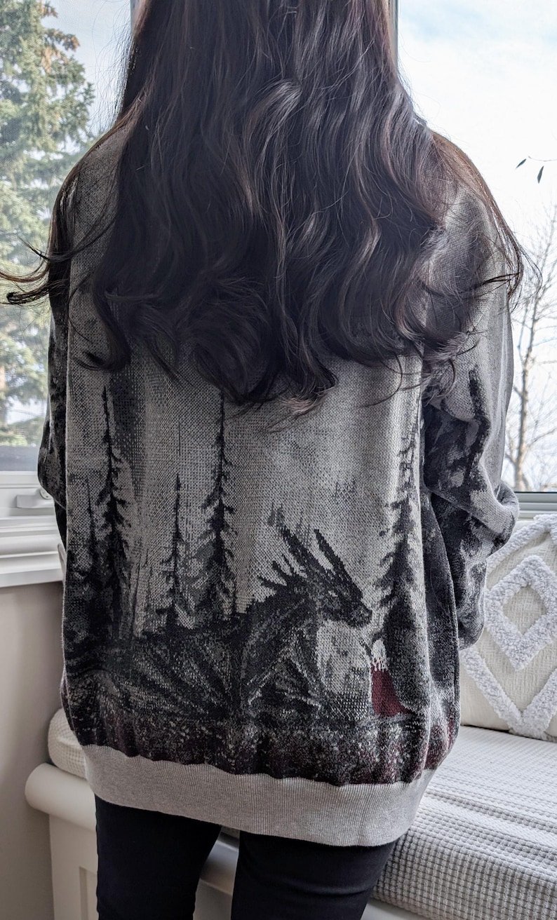 May include: A gray sweater featuring a forest scene with a dragon and a figure in red. The design is in shades of gray and black, with a gray hem and cuffs. The sweater is worn by a person with long, dark hair.