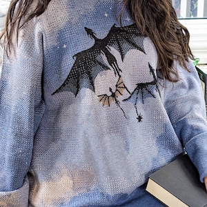 K&ouml;nnte beinhalten: Ein lang&auml;rmeliger blauer Kapuzenpullover mit einem Strickmuster aus n&auml;chtlichem Himmel mit Wolken und drei schwarzen fliegenden Drachensilhouetten. Das Kleidungsst&uuml;ck hat einen gerippten Saum und B&uuml;ndchen, eine Kapuze und eine Textur.