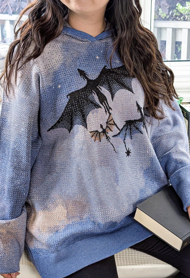 K&ouml;nnte beinhalten: Ein lang&auml;rmeliger blauer Kapuzenpullover mit einem Strickmuster aus n&auml;chtlichem Himmel mit Wolken und drei schwarzen fliegenden Drachensilhouetten. Das Kleidungsst&uuml;ck hat einen gerippten Saum und B&uuml;ndchen, eine Kapuze und eine Textur.