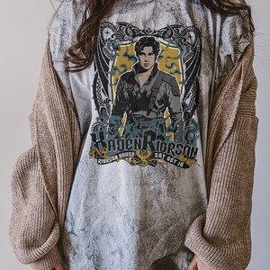 May include: A grey t-shirt with a faded, distressed look. The shirt features a graphic of a man with a sword, surrounded by a dragon design. The text "Riorsson House" and "Sat Oct 10" is printed below the graphic.