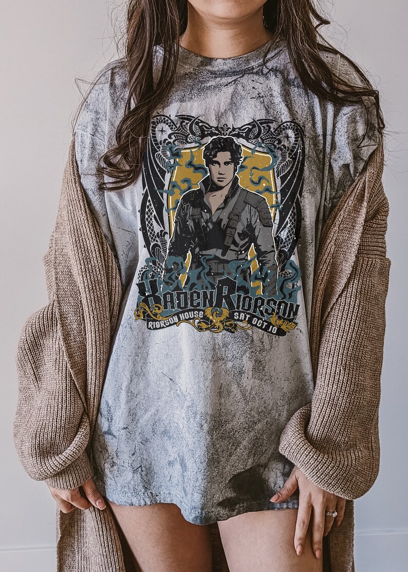 May include: A grey t-shirt with a faded, distressed look. The shirt features a graphic of a man with a sword, surrounded by a dragon design. The text "Riorsson House" and "Sat Oct 10" is printed below the graphic.