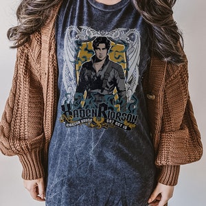 May include: A dark grey t-shirt with a faded vintage look featuring a graphic of a man with dark hair and a black shirt. The graphic is surrounded by a gold and white design with the text "Kaden Riorson Riorson House Sat Oct 10".
