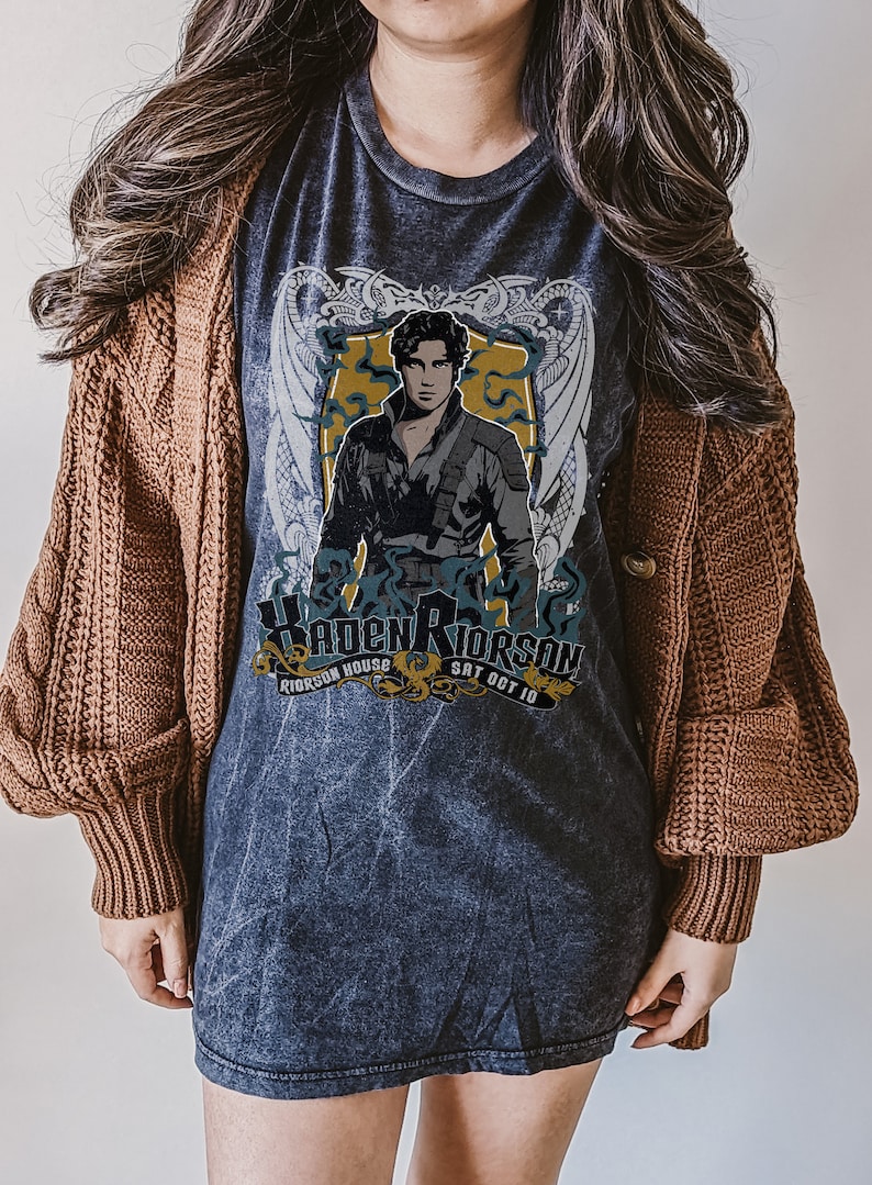 May include: A dark grey t-shirt with a faded vintage look featuring a graphic of a man with dark hair and a black shirt. The graphic is surrounded by a gold and white design with the text "Kaden Riorson Riorson House Sat Oct 10".