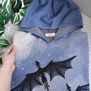 K&ouml;nnte beinhalten: Ein blauer Strick-Kapuzenpullover mit einem grafischen Muster aus drei schwarzen Drachen, die vor einem hellblauen und cremefarbenen Himmel mit kleinen wei&szlig;en Sternen fliegen. Die Innenseite der Kapuze ist braun und ein wei&szlig;es Etikett zeigt Narrative Threads.