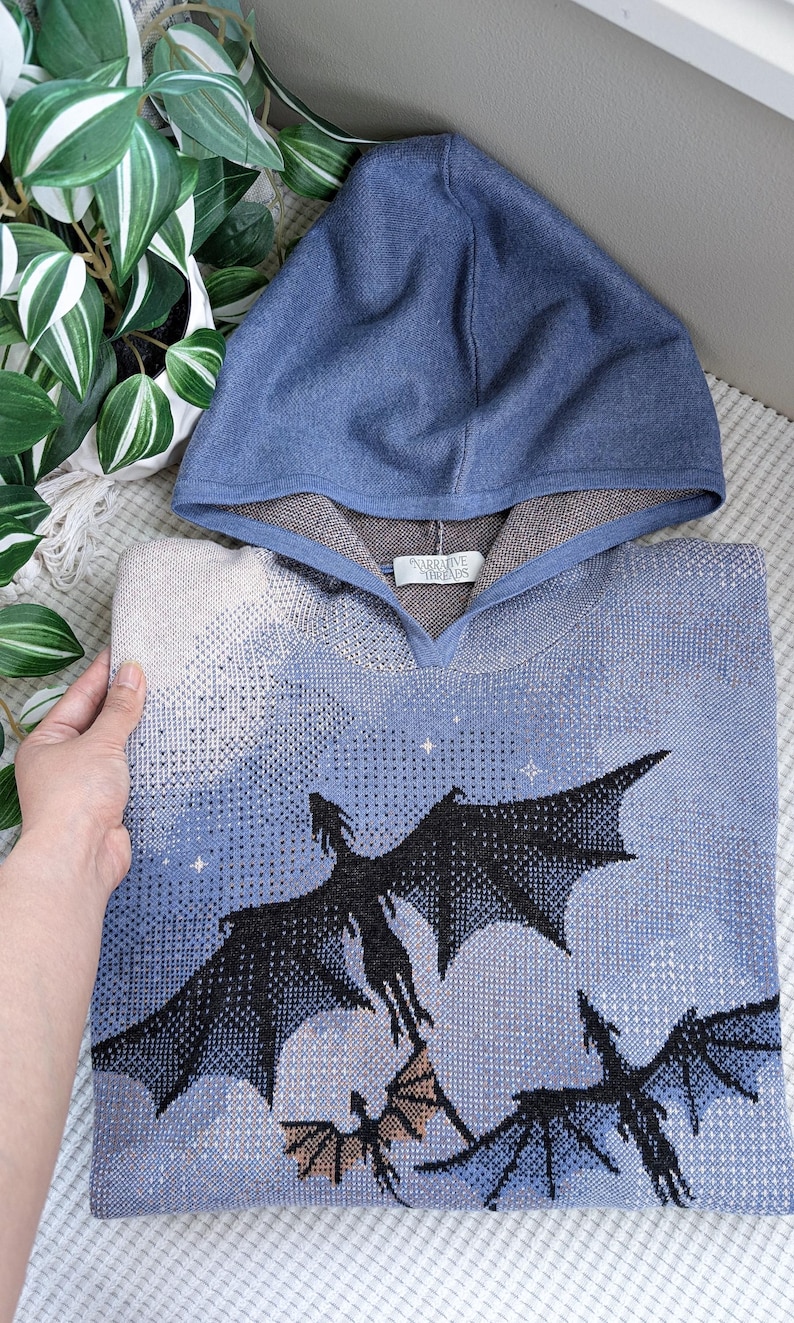 K&ouml;nnte beinhalten: Ein blauer Strick-Kapuzenpullover mit einem grafischen Muster aus drei schwarzen Drachen, die vor einem hellblauen und cremefarbenen Himmel mit kleinen wei&szlig;en Sternen fliegen. Die Innenseite der Kapuze ist braun und ein wei&szlig;es Etikett zeigt Narrative Threads.
