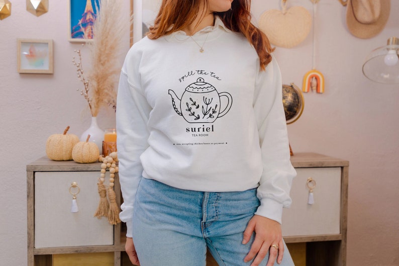 May include: White sweatshirt with a black and white illustration of a teapot with the text "spill the tea" and "suriel tea room".