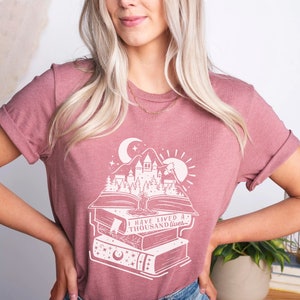 May include: A mauve t-shirt with a white graphic design of a castle and forest scene on top of a stack of books. The text on the shirt reads "I have lived a thousand lives."
